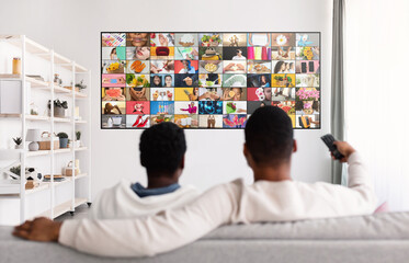 Obraz premium Two men sit on a white couch facing a large flat-screen television with a grid of various images on the screen. One man is holding a remote control in his right hand.
