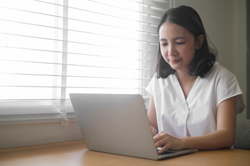 Woman working from home on a laptop, sitting by a window with blinds, focused and productive in a casual white shirt.