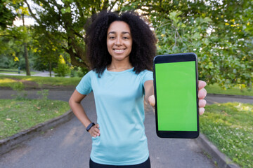 An African American female athlete smiling confidently as she holds a smartphone with a green screen in an outdoor fitness class setting.