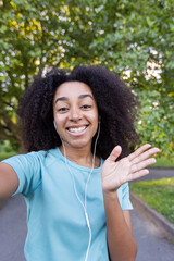 Cheerful African American female athlete waves hello during an outdoor fitness class while using a phone app for a video call. She is wearing workout clothes and earbuds