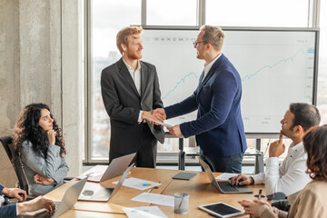 Two businessmen in suits are shaking hands in a modern office, while other colleagues look on. Large screen behind them displaying a graph, and laptops and other work materials are on the table.