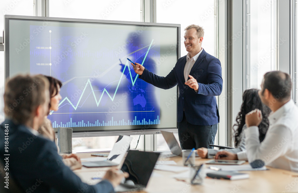Poster A businessman stands at the front of a meeting room, pointing at a large screen displaying a financial graph. He is presenting data to his colleagues, who are seated around a conference table.