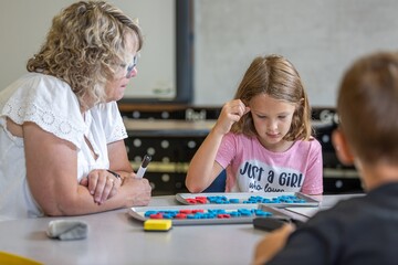 Naklejka premium A girl in a pink shirt is sitting at a table with a woman and a boy. The girl is learning with blocks and the woman is watching her. Scene is playful and lighthearted