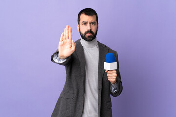 Adult reporter man with beard holding a microphone over isolated purple background making stop...