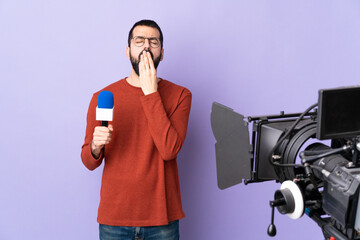 Reporter man holding a microphone and reporting news over isolated purple background yawning and covering wide open mouth with hand