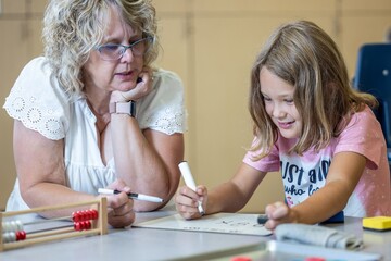 A teacher is helping a young girl with her math homework. The girl is wearing a pink shirt and is using a white marker to write on a small white board. The teacher is sitting next to her