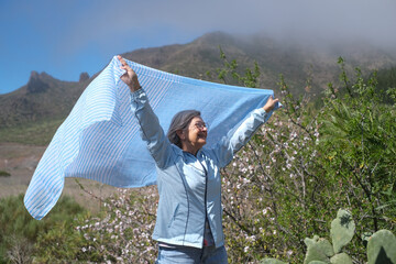 Smiling senior woman with gray hair waving a scarf in outdoors in a sunny day near flowering almond...
