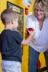 A young boy is holding a red apple and giving it to his teacher. The teacher is wearing glasses and has a white shirt on. The scene appears to be a casual