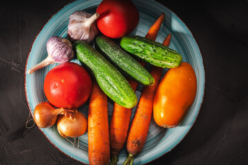 Summer vegetables on a turquoise plate on a dark gray background: tomatoes, cucumbers, carrots, garlic, onions. Copy space.