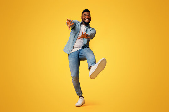 Funny african american guy dancing and extending his hands to camera over yellow studio background, low-angle view