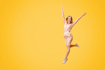 Obraz premium Freedom Of Youth. Portrait of carefree teen girl in summer dress jumping in the air with raised arms over yellow studio background, copy space