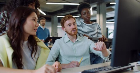Camera view of Caucasian male sitting at desk and explaining something to new interns or managers. Man pointing with hand at monitor and teaching newcomers about important company lessons. Teamwork.