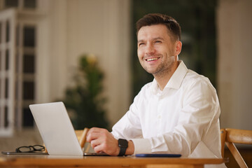 Smiling man in a white shirt working on a laptop at a cozy indoor setting, exuding positivity and...