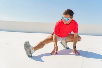 A man in a bright red shirt stretching outdoors under a clear blue sky, wearing reflective sunglasses, focused on his warm-up.