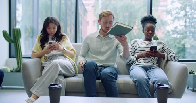 Camera moving closer to multi-ethnic group of strangers sitting on grey couch. Man in middle taking notebook from coffee table while waiting for something. Girls using mobile devices. Busy people.