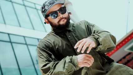 Asian hipster looking at camera while dancing at sky scrapper. Attractive handsome hipster moving his hands to hip hop music at modern mall with low angle camera. Outdoor sport 2024. Hiphop.