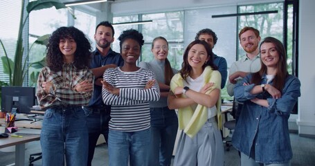 Front view of multi-ethnic group of people standing next to each other. Everyone crossing their hands and smiling while looking directly at camera. Posing for team photo. Office in background.