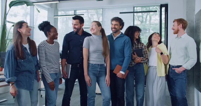 Team of friendly coworkers standing next to each other and looking directly at camera while smiling. Wearing casual outfits. Modern office in background. Workers holding notebooks and gadgets.