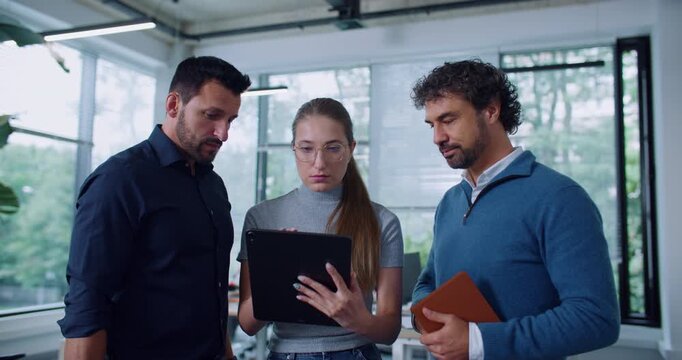 Caucasian group of people having discussing in middle of office building. Woman with glasses holding tablet while listening to man. Male pointing at screen and explaining something to colleagues.