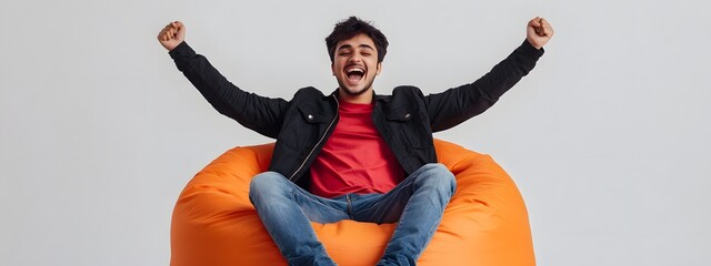 A young man sitting on an orange bean bag chair, wearing jeans and black jacket with red t-shirt underneath. He is excitedly celebrating his success in front of the camera against white background