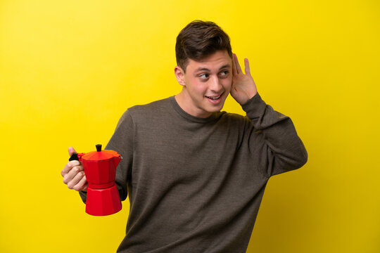 Young Brazilian man holding coffee pot isolated on yellow background listening to something by putting hand on the ear