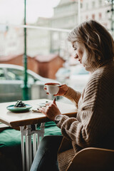 A woman is sitting at a table with a cup of coffee in front of her. She is wearing a brown sweater and is looking at her phone. The scene is set in a cafe, with a car visible in the background