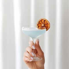 A close-up shot of a hand holding a light blue cocktail in a delicate coupe glass with a long, thin stem. A slice of dried orange is elegantly placed on the rim of the glass.