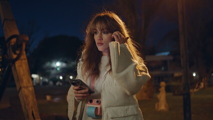 Fototapeta premium Serious lady looking cellphone standing in night park closeup. Worried girl