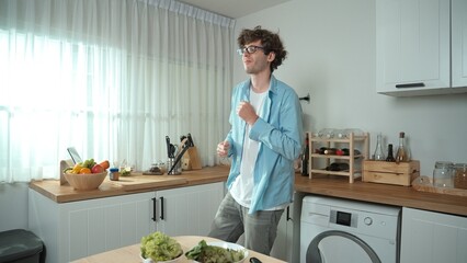 Happy father move to music while cooking food and prepare lunch at modern kitchen. Active dad dancing to song while making healthy food or salad surrounded with vegetable and kitchenware. Pedagogy.