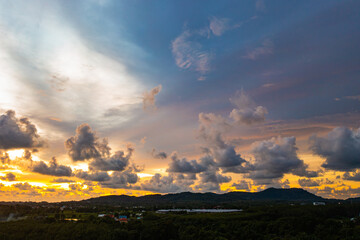 A breathtaking aerial view capturing a vibrant sunset over the Phuket countryside. The sky is ablaze with rich hues of yellow, orange, pink, and purple, while dramatic clouds float above the city