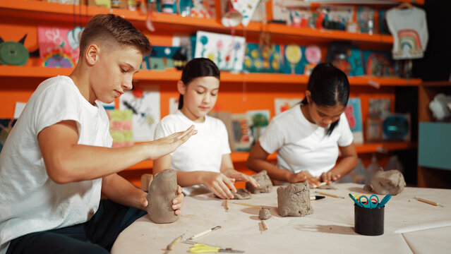 Caucasian smart teenager modeling clay while craving glass of clay at art lesson. Group of multicultural happy student working at dough in pottery workshop. Creative activity concept. Edification.
