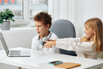 Happy Caucasian siblings studying together at home The boy and girl are sitting at a table in the living room, using their laptops for online elearning They are wearing headphones and concentrating on