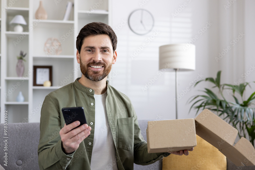 Wall mural man smiling holding phone and open cardboard box in living room. concept of online shopping, deliver