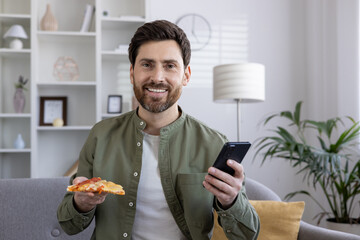 Man holding slice of pizza and using phone in living room. Relaxed man smiling, enjoying casual moment at home, multitasking with food and technology.