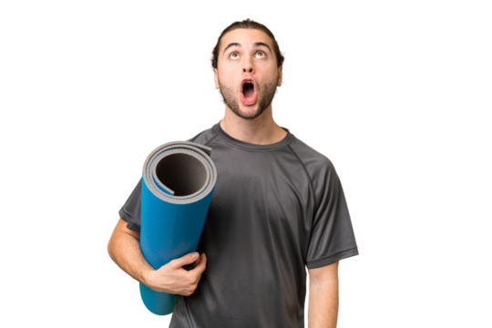 Young sport man going to yoga classes while holding a mat over isolated background looking up and with surprised expression