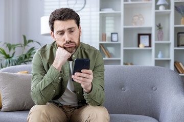 Thoughtful man sitting on sofa looking at smartphone at home. Pensive expression, appears deep in...