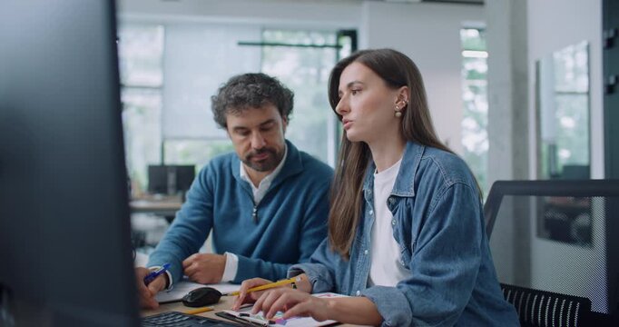 Pretty Caucasian woman with her colleague working together at computer in office. People speaking about business project. Looking with focus at display while sitting at table in office. Collaboration.