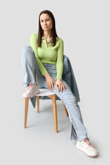 Beautiful young woman in denim clothes sitting on chair against grey background