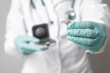 Female doctor in rubber gloves holding hearing aid against grey wall, closeup