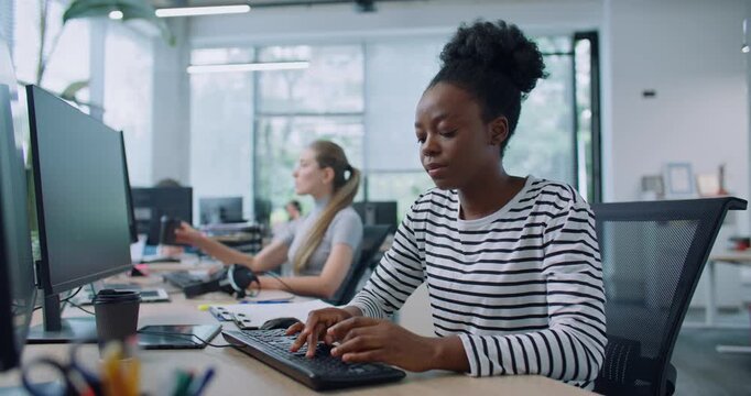 Zooming in on pretty woman with casual clothes working at computer in office. Distracted by camera from work and joyfully smiling. In background people sitting at their workplaces. Job concept.
