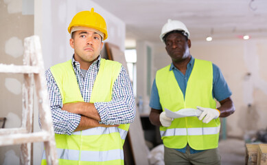 Portrait of confused caucasian man builder standing in construction site. His boss, african-american man, standing in background with documents.