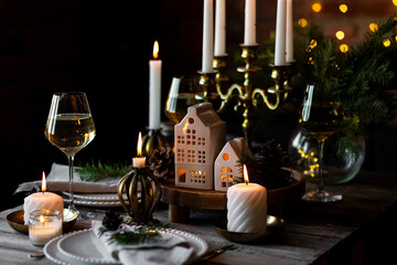 Luxury elegant Christmas dinner table setting with old vintage golden chandelier, candles, fir tree branches, wooden furniture. Porcelain houses as centrepiece, garland with lights on background 