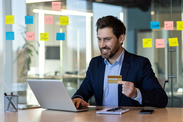 Smiling businessman using laptop and holding credit card. Man working in office, making online payment with phone on desk. E-commerce, business, technology.