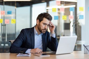 Businessman in suit looking stressed while working on laptop in modern office with sticky notes on glass wall. Concept of workplace stress, pressure, and productivity