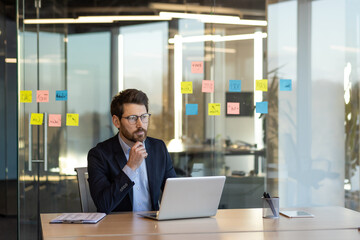 Professional businessman using laptop in modern office setting with glass walls and colorful sticky...