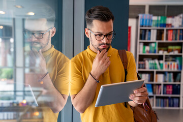 A male student, clad in a yellow tee, thoughtfully uses his digital tablet while leaning against...