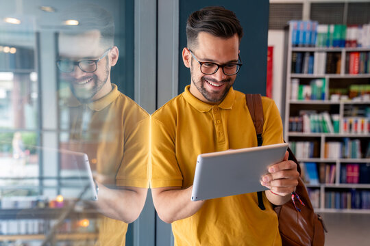 A joyful Caucasian male student, dressed in a yellow shirt, uses a digital tablet while leaning against a library window, reflecting his studious endeavor.