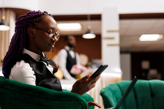 Young African American woman hotel employee looking at smartphone screen while sitting at lobby with laptop, using mobile application tracking booking flow. Inventory software in hospitality industry