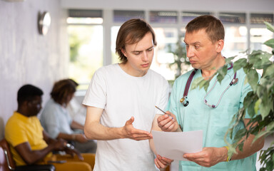 Obraz premium Serious middle-aged male doctor discussing medical document with concerned patient standing in waiting room