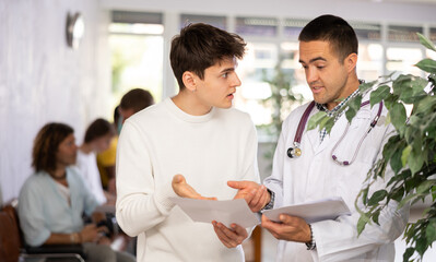 Young male doctor and patient discussing medical document standing in waiting room